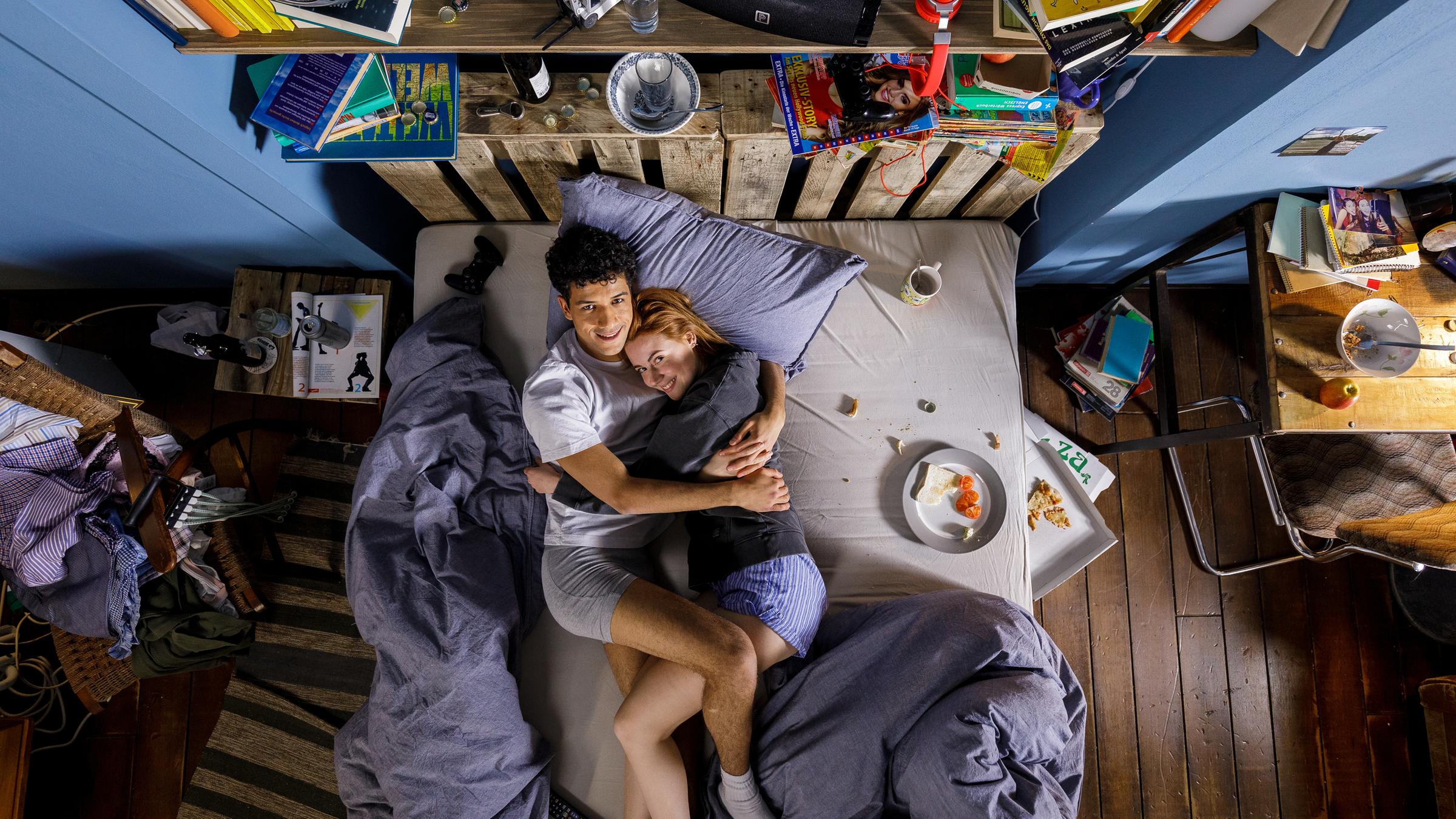 "Ich dich auch! - Key Visual": Caro (Meriel Hinsching) und Yannik (Rojan Juan Barani) liegen kuschelnd auf dem Bett. Beide sind angezogen, lächeln glücklich in die Kamera. Auf dem Bett und daneben stehen Teller und Tassen mit Essensresten, das Zimmer wirkt unaufgeräumt.