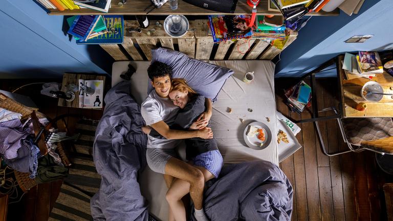 "Ich dich auch! - Key Visual": Caro (Meriel Hinsching) und Yannik (Rojan Juan Barani) liegen kuschelnd auf dem Bett. Beide sind angezogen, lächeln glücklich in die Kamera. Auf dem Bett und daneben stehen Teller und Tassen mit Essensresten, das Zimmer wirkt unaufgeräumt.