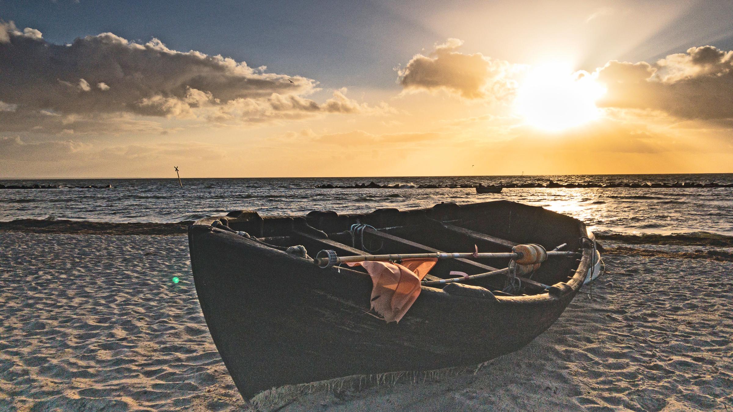 "Aussterbende Art": Abendstimmung am Strand. Ein hölzernes Fischerboot liegt im Sand. Im Hintergrund das Meer und die untergehende Sonne.