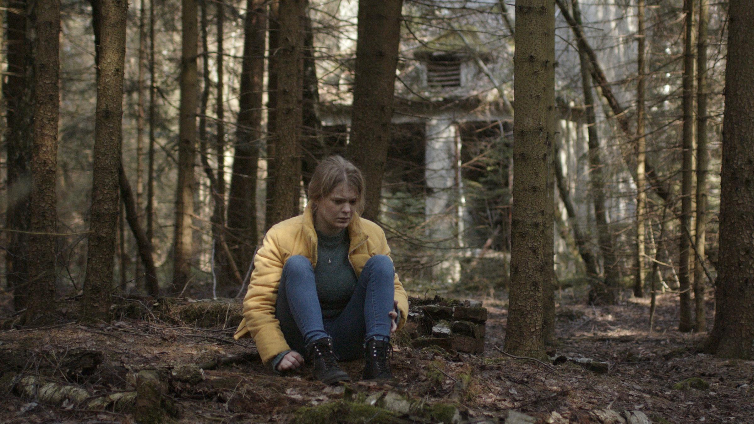 "Schlaf": Mona (Gro Swantje Kohlhof) sitzt auf dem Waldboden und starrt vor sich hin. Im Hintergrund eine leerstehendes Haus.