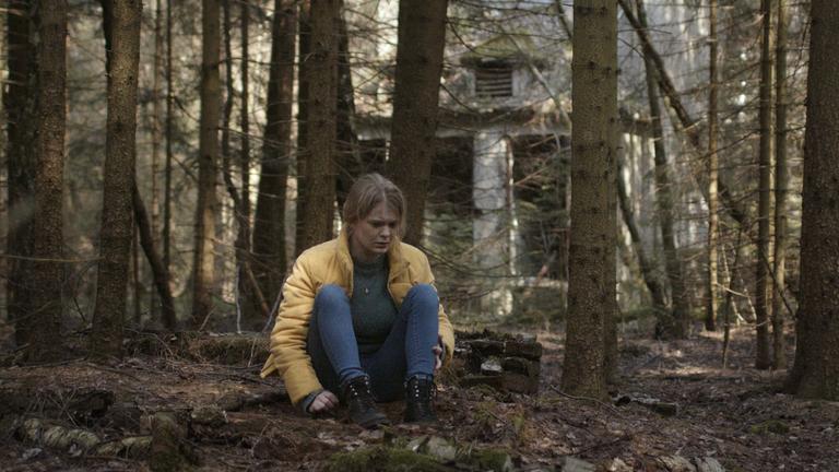"Schlaf": Mona (Gro Swantje Kohlhof) sitzt auf dem Waldboden und starrt vor sich hin. Im Hintergrund eine leerstehendes Haus.