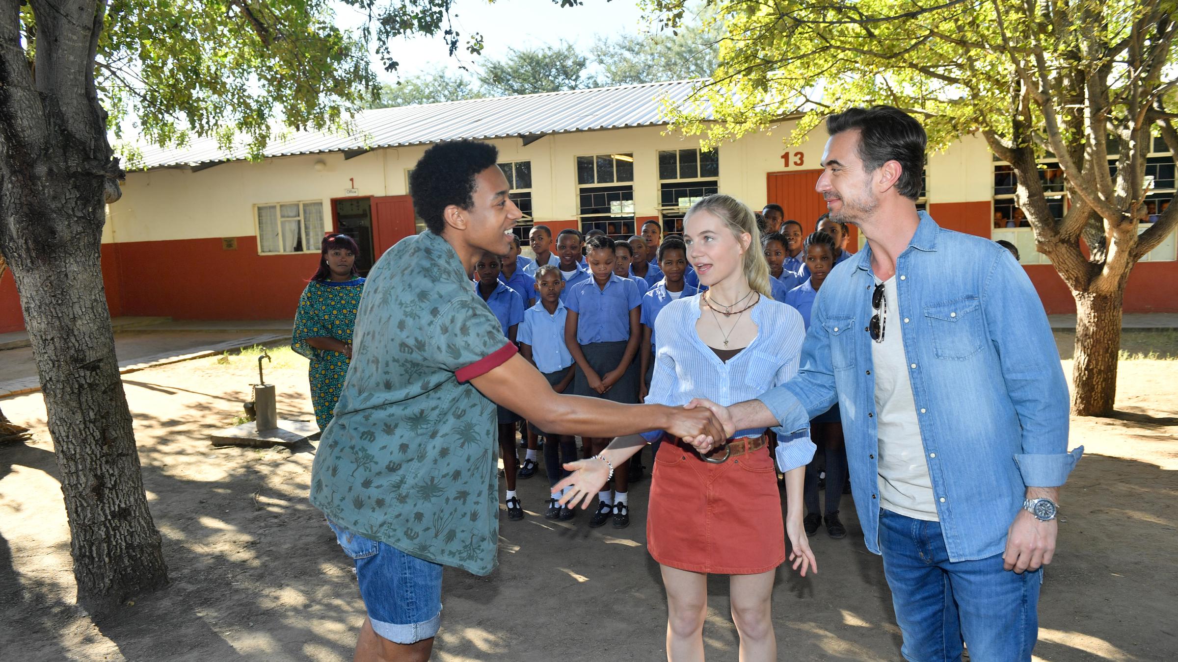 "Das Traumschiff: Namibia": Vor einem Schulgebäude steht Max Parger (Florian Silbereisen) und reicht Tayo Balewa (Tidiane Jemay Diallo) lächelnd die Hand. Bei ihnen steht redend Leonie Parger (Louise Sophie Arnold). Im Hintergrund stehen Kinder in Schuluniformen.