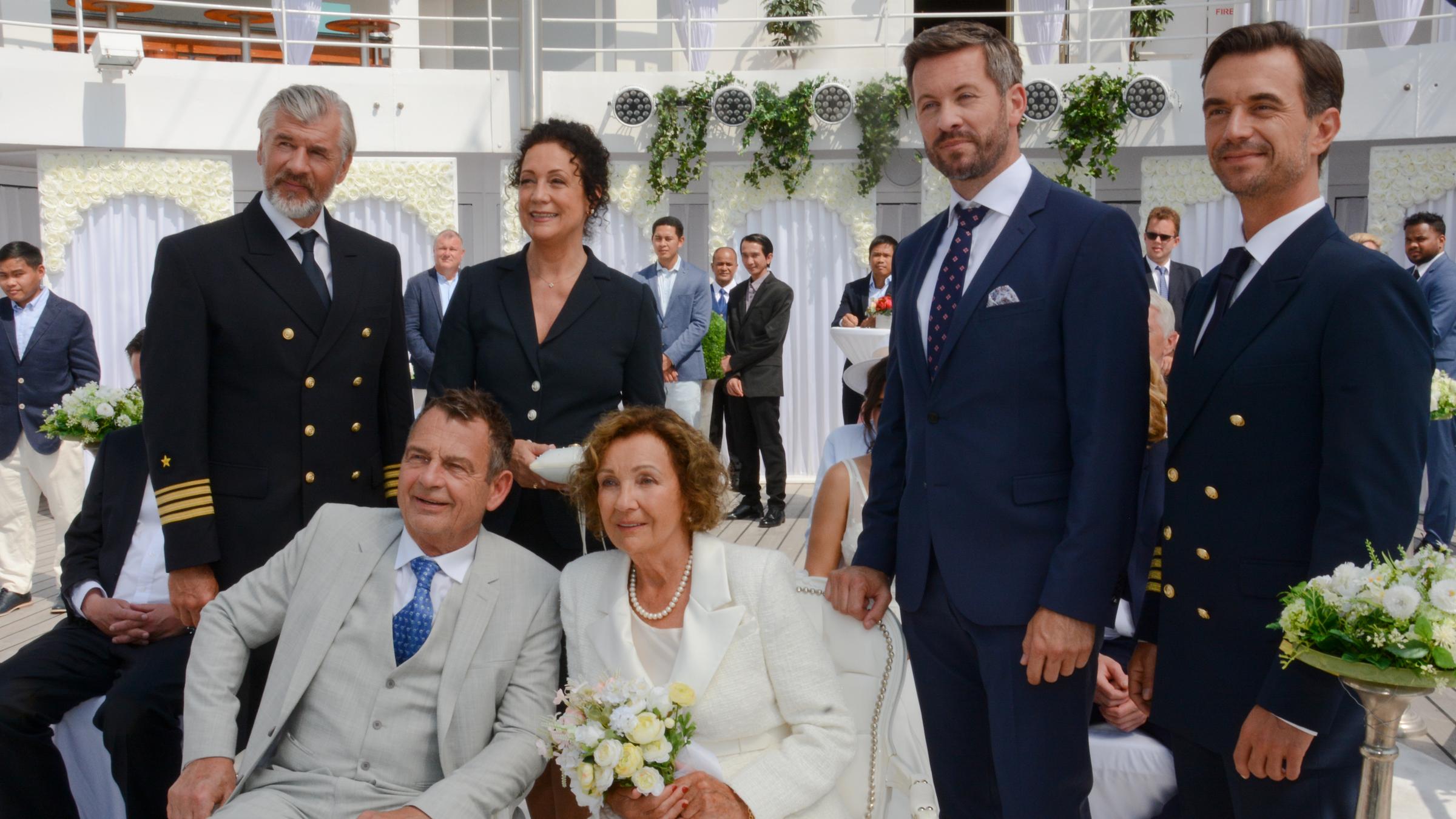 "Kreuzfahrt ins Glück - Hochzeitsreise nach Kreta": Max Parger (Florian Silbereisen), Hanna Liebhold (Barbara Wussow), Martin Grimm (Daniel Morgenroth) und Tom Cramer (Jan Hartmann) stehen lächelnd hinter Kerstin Cramer (Gila von Weitershausen) und Achim Vormann (Krystian Martinek), die in ihrem Hochzeitsoutfit auf Stühlen sitzen.