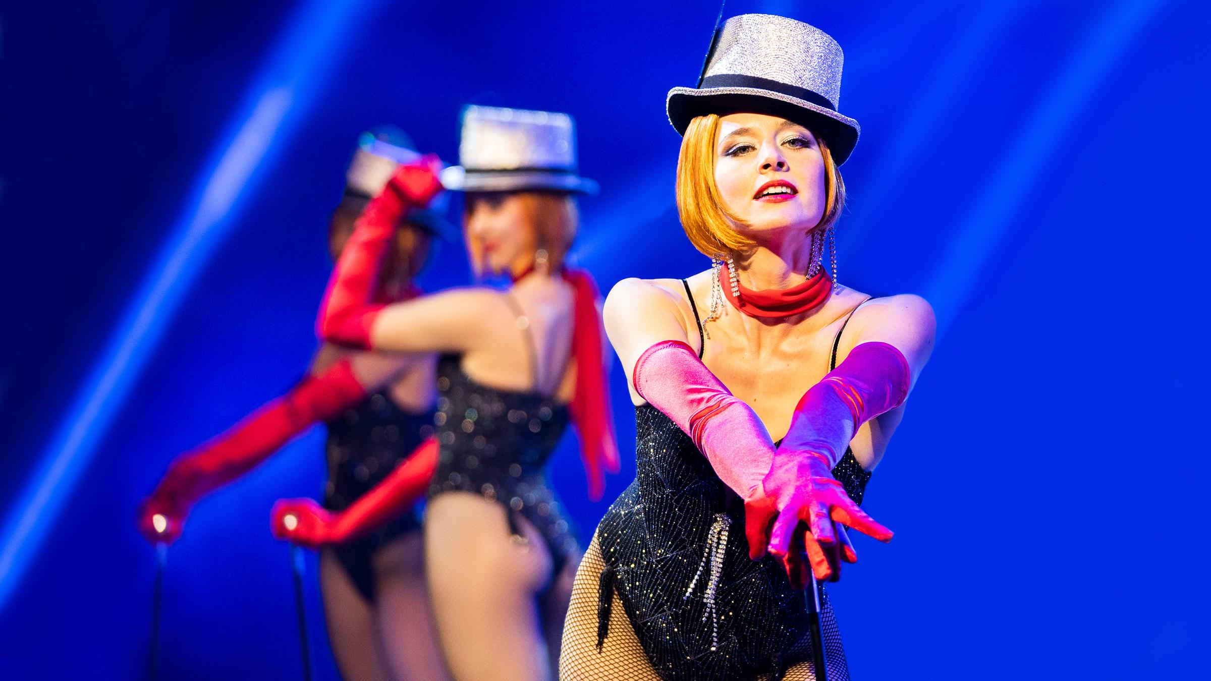 „Der Palast“: Die Tänzerin Chris/Marlene (Svenja Jung) posiert mit einem Tanzstab. Sie trägt einen schwarzen Body, rote Handschuhe und einen silbernen Hut. Im Hintergrund posieren zwei weitere Tänzerinnen.