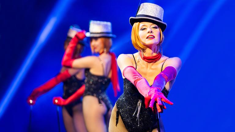 „Der Palast“: Die Tänzerin Chris/Marlene (Svenja Jung) posiert mit einem Tanzstab. Sie trägt einen schwarzen Body, rote Handschuhe und einen silbernen Hut. Im Hintergrund posieren zwei weitere Tänzerinnen.