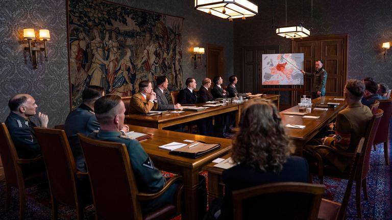 "Die Wannseekonferenz": Vor dem U-förmigen Besprechungstisch  steht Adolf Eichmann (Johannes Allmayer) vor einer Tafel, auf der die Europakarte zu sehen ist. In seiner rechten Hand hält er einen Zeigestock und zeigt auf die Karte. Die anderen Teilnehmer der Besprechung sitzen am Tisch und schauen  konzentriert auf ihn.
