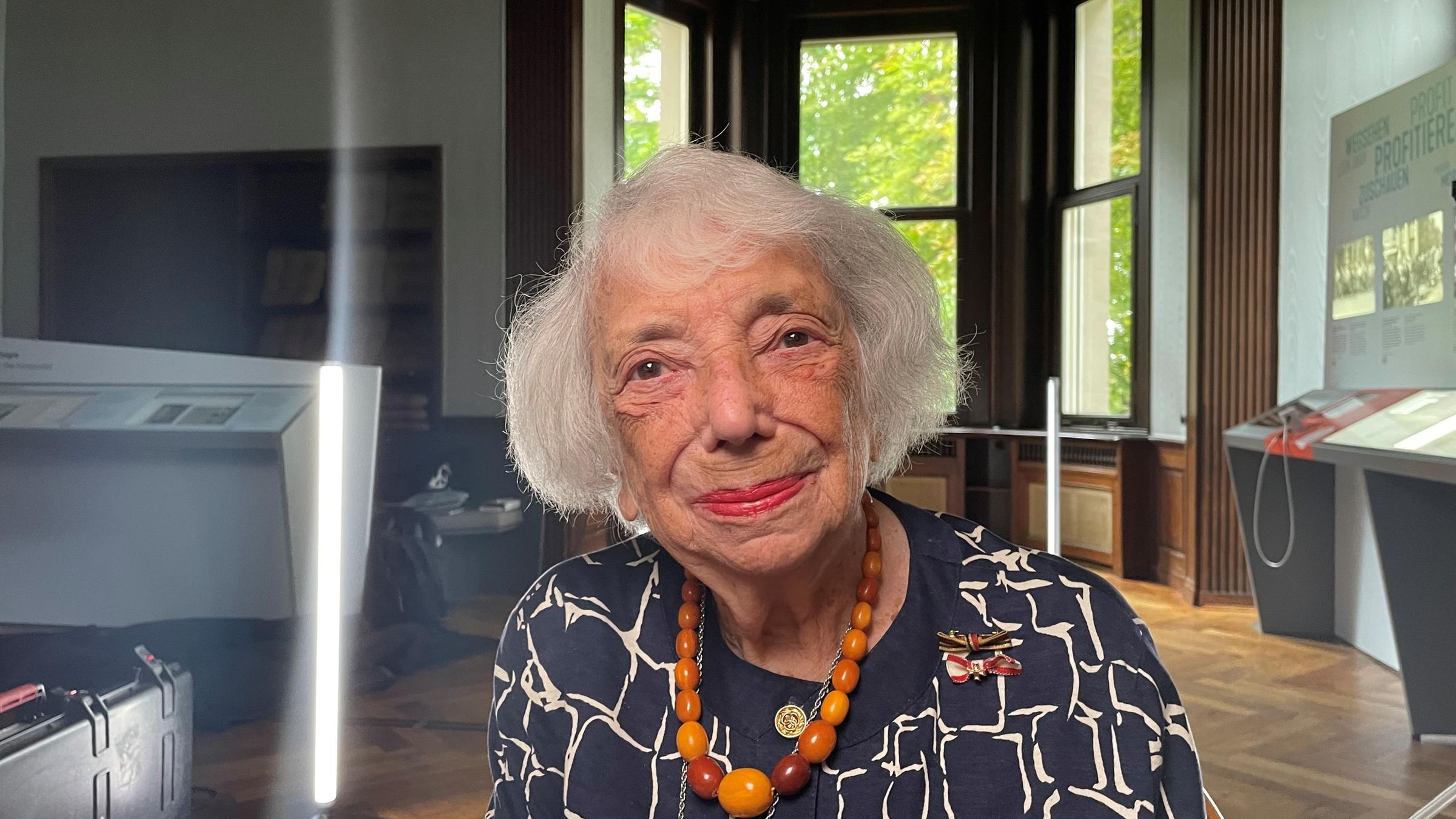 "Die Wannseekonferenz - Begleitdoku": Eine alte Dame sitzt in einem modern ausgestatteten hohen Raum der historischen Ursprung hat.