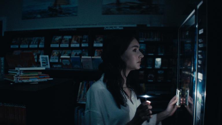"Sløborn - Alles anders": In einer dunklen Buchhandlung inspiziert Evelin (Emily Kusche) den Inhalt eines Snackautomaten.
