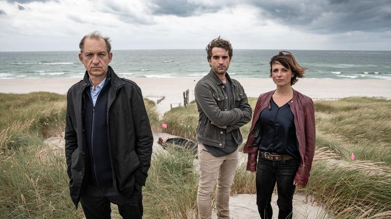 "Nord Nord Mord - Sievers und das mörderische Türkis": Carl Sievers (Peter Heinrich Brix), Hinnerk Feldmann (Oliver Wnuk) und Ina Behrendsen (Julia Brendler) stehen in den Dünen. Hinter ihnen liegt im Sand ein lebloser braunhaariger Mann, der mit einem grünen Anorak bekleidet ist. Es ist Harald Piontek (Mischa Latwesen).  Es ist sehr windig. Im Hintergrund ist die Brandung der Nordsee und Strand zu sehen.