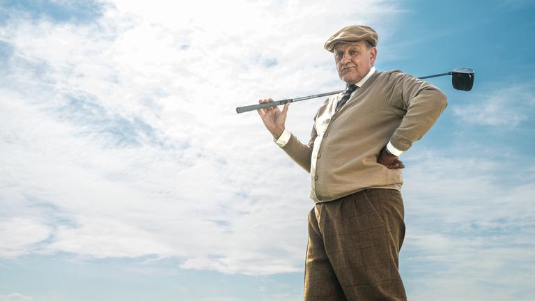 "Wilsberg - Gene lügen nicht": Wilsberg (Leonard Lansink) steht auf dem gepflegten Rasen eines Golfplatzes, hat einen Golfschläger lässig über seine Schulter gelegt und schaut auf einen Golfball, der im Vordergrund liegt. Er trägt eine Schiebermütze, Hemd und Krawatte unter einer braunen Strickjacke, eine knielange Cordhose und weiße Kniestrümpfe. Im Hintergrund steht das elegante Clubhaus des Golfplatzes.