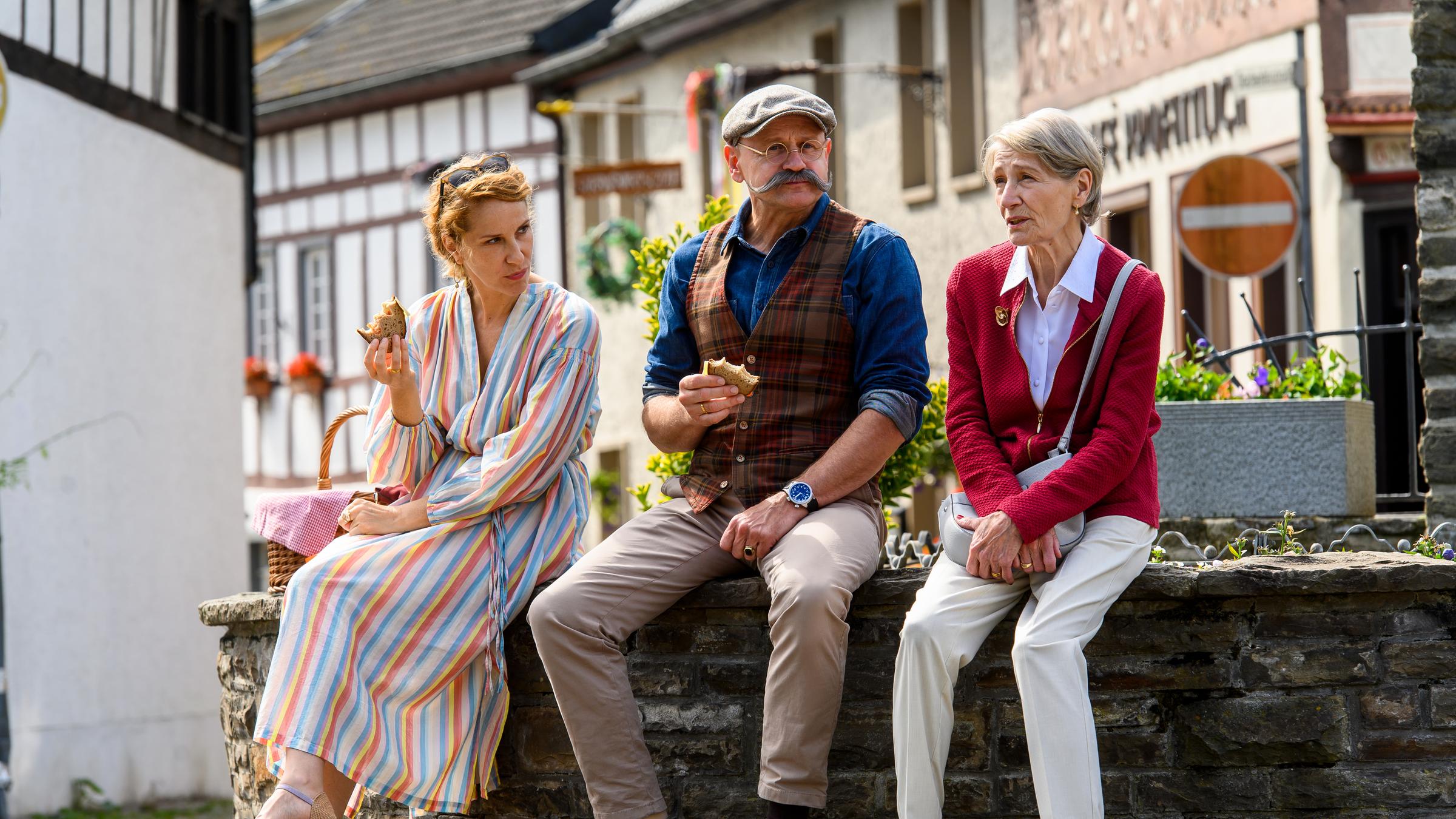 "Horst Lichter - Keine Zeit für Arschlöcher": Nada Lichter (Chiara Schoras), Horst Lichter (Oliver Stokowski) und Margret Lichter (Barbara Nüsse) sitzen in einer Fachwerkstadt an einem Brunnen. Nada und Horst haben ein Vesperbrot in der Hand. Margret hält sich an ihrer Handtasche fest.