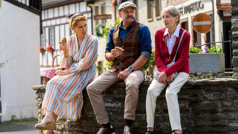 "Horst Lichter - Keine Zeit für Arschlöcher": Nada Lichter (Chiara Schoras), Horst Lichter (Oliver Stokowski) und Margret Lichter (Barbara Nüsse) sitzen in einer Fachwerkstadt an einem Brunnen. Nada und Horst haben ein Vesperbrot in der Hand. Margret hält sich an ihrer Handtasche fest.
