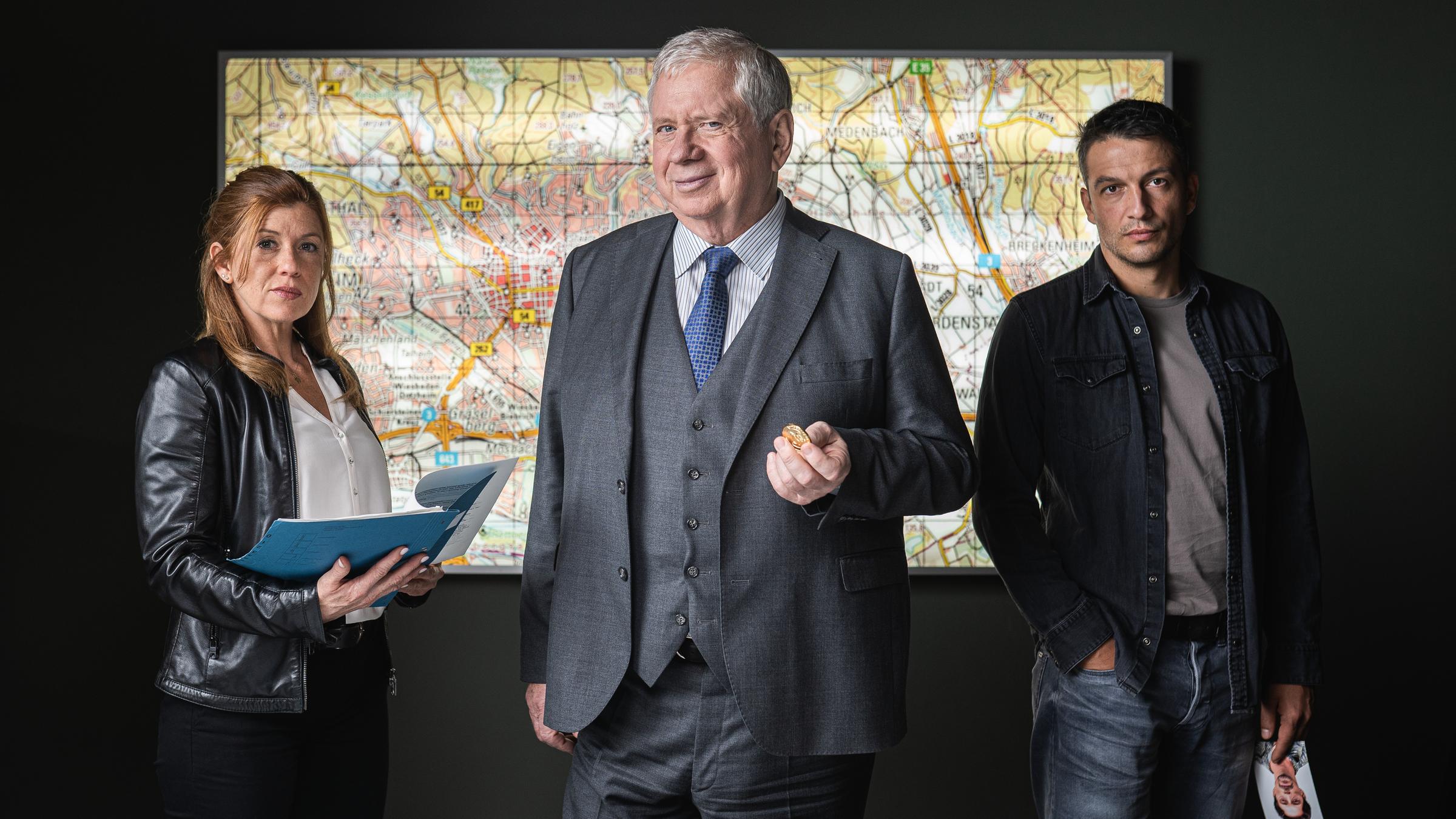 "Der Staatsanwalt - Tödliches Dickicht": Oberstaatsanwalt Bernd Reuther (Rainer Hunold), Hauptkommissarin Kerstin Klar (Fiona Coors) und Oberkommissar Max Fischer (Max Hemmersdorfer) stehen hinter einem Schreibtisch und blicken entschlossen in die Kamera. Reuther hält eine Goldmünze in seiner linken Hand. Auf dem Schreibtisch liegen diverse Unterlagen, im Hintergrund an der Wand ist ein Stadtplan zu sehen.
