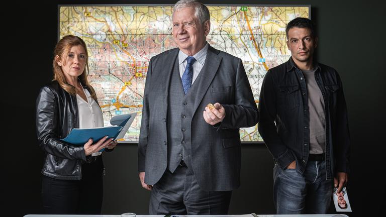"Der Staatsanwalt - Tödliches Dickicht": Oberstaatsanwalt Bernd Reuther (Rainer Hunold), Hauptkommissarin Kerstin Klar (Fiona Coors) und Oberkommissar Max Fischer (Max Hemmersdorfer) stehen hinter einem Schreibtisch und blicken entschlossen in die Kamera. Reuther hält eine Goldmünze in seiner linken Hand. Auf dem Schreibtisch liegen diverse Unterlagen, im Hintergrund an der Wand ist ein Stadtplan zu sehen.