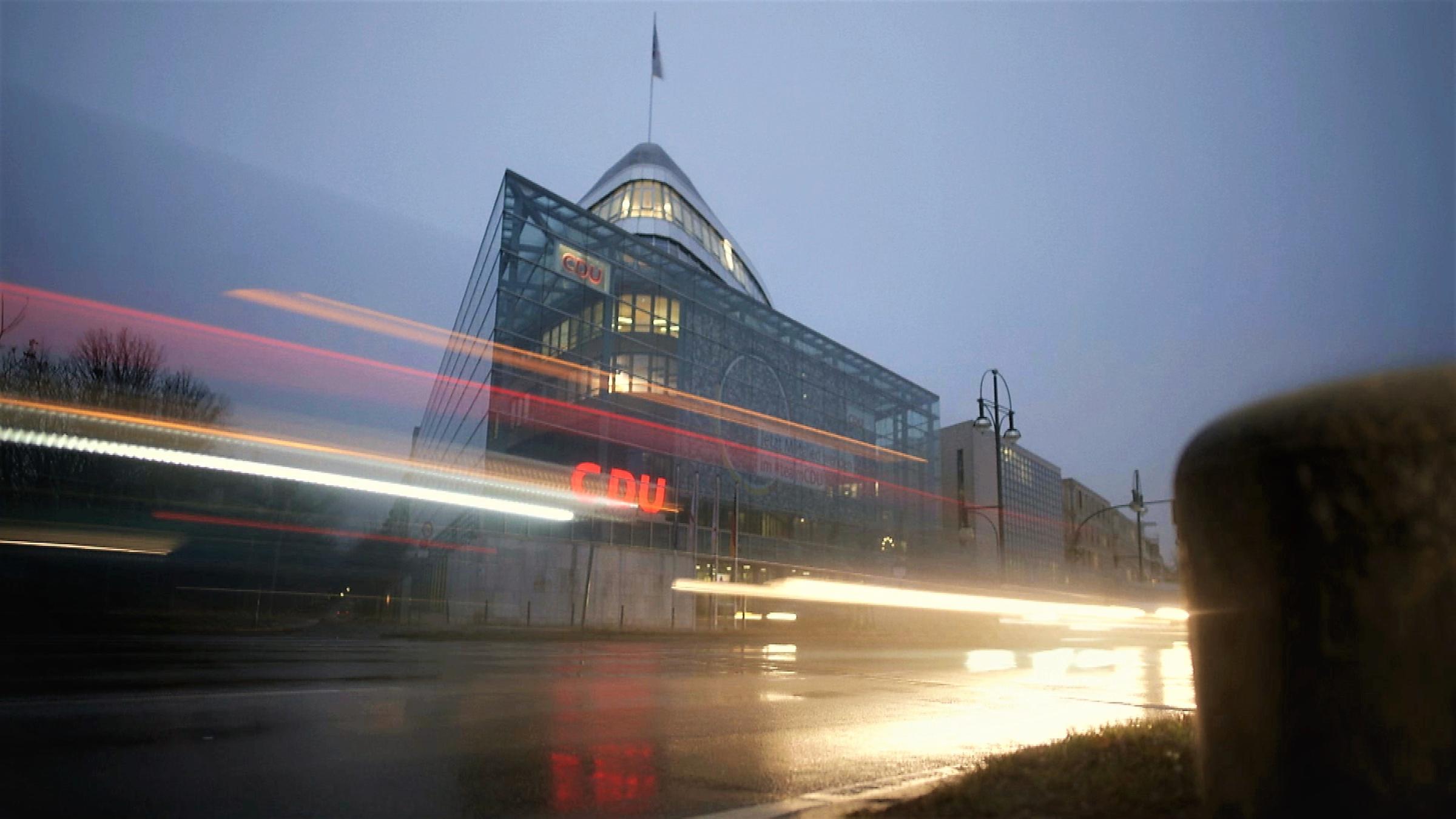  "Verstörte Volkspartei - Eine Reise durch die CDU": Blick auf das Konrad-Adenauer-Haus in der Dämmerung. Im Vordergrund eine regennasse Straße mit Scheinwerfer-Reflektionen.