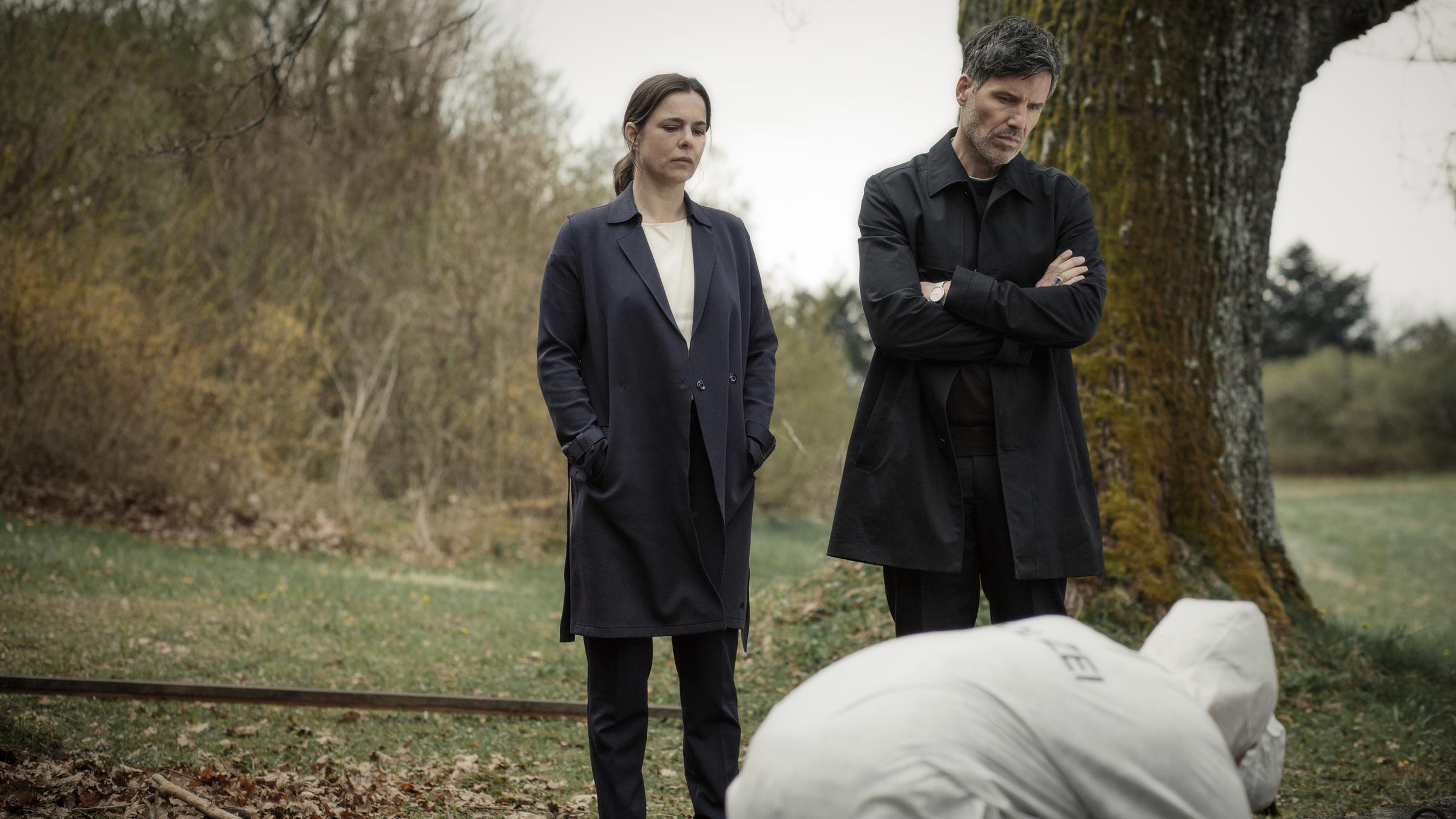 "Muttertag (1) - Ein Taunuskrimi": In einem parkähnlichen Gelände stehen Pia Sander (Annika Kuhl) und Oliver von Bodenstein (Tim Bergmann) und blicken auf zwei vor ihnen am Boden arbeitende KollegInnen im weißen Schutzanzug. Ein mächtiger Baum steht hinter Oliver, trockenes Laub liegt neben Pia am Boden. Im Hintergrund stehen Büsche.