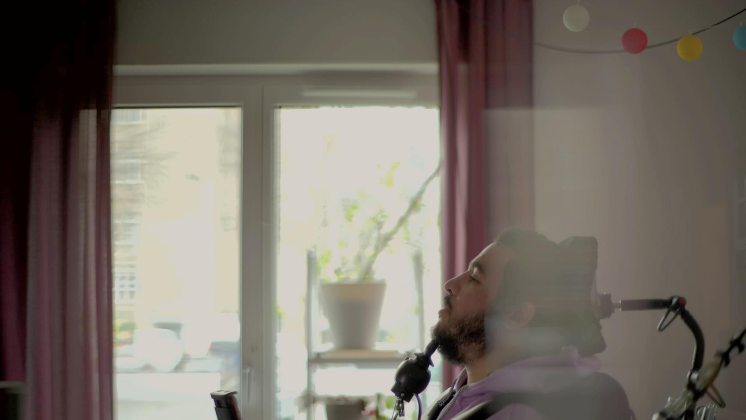 "37°: Der Tod- Die beste Entscheidung meines Lebens": Noah Berge , ein junger Mann, sitzt im Rollstuhl. Man sieht ihn von der Seite. Im Hintergrund sieht man ein Fenster, an dem rote Gardinen hängen und ein Regal mit einer Pflanze darauf.