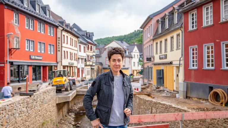 "Die Flut - Zwischen Wut und Mut: Ein Jahr nach dem Hochwasser": Im Bildvordergrund steht die ZDF-Reporterin Dunja Hayali auf einer Behelfsbrücke in der Fußgängerzone von Bad Münstereifel. Im Hintergrund sieht man Aufbauarbeiten am Flusslauf entlang der Erft. Sie fließt durch die historische Altstadt.