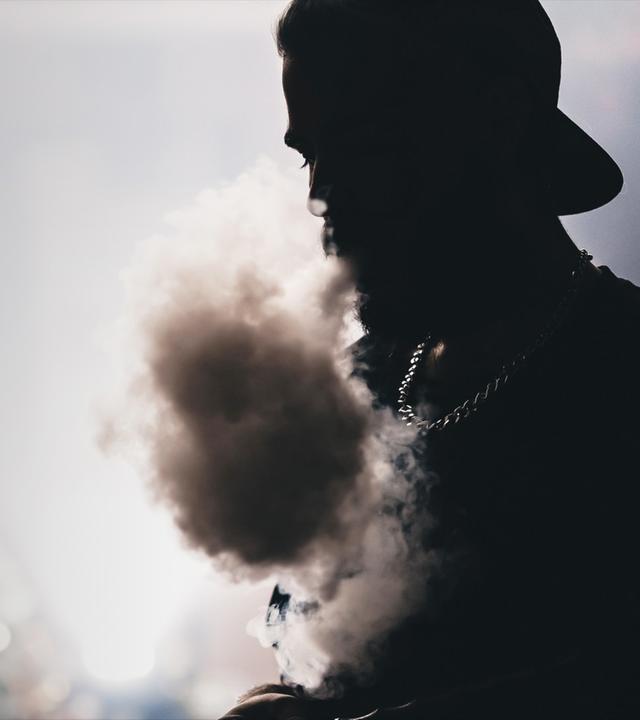 "ZDFzeit: Kriminelle Clans - Geld, Macht und Ehre": Die Silhouette eines Mannes, dessen Kette im schwachen Licht glänzt. Er trägt eine Kappe und stößt Shisha-Rauch aus. Der Hintergrund ist unscharf.