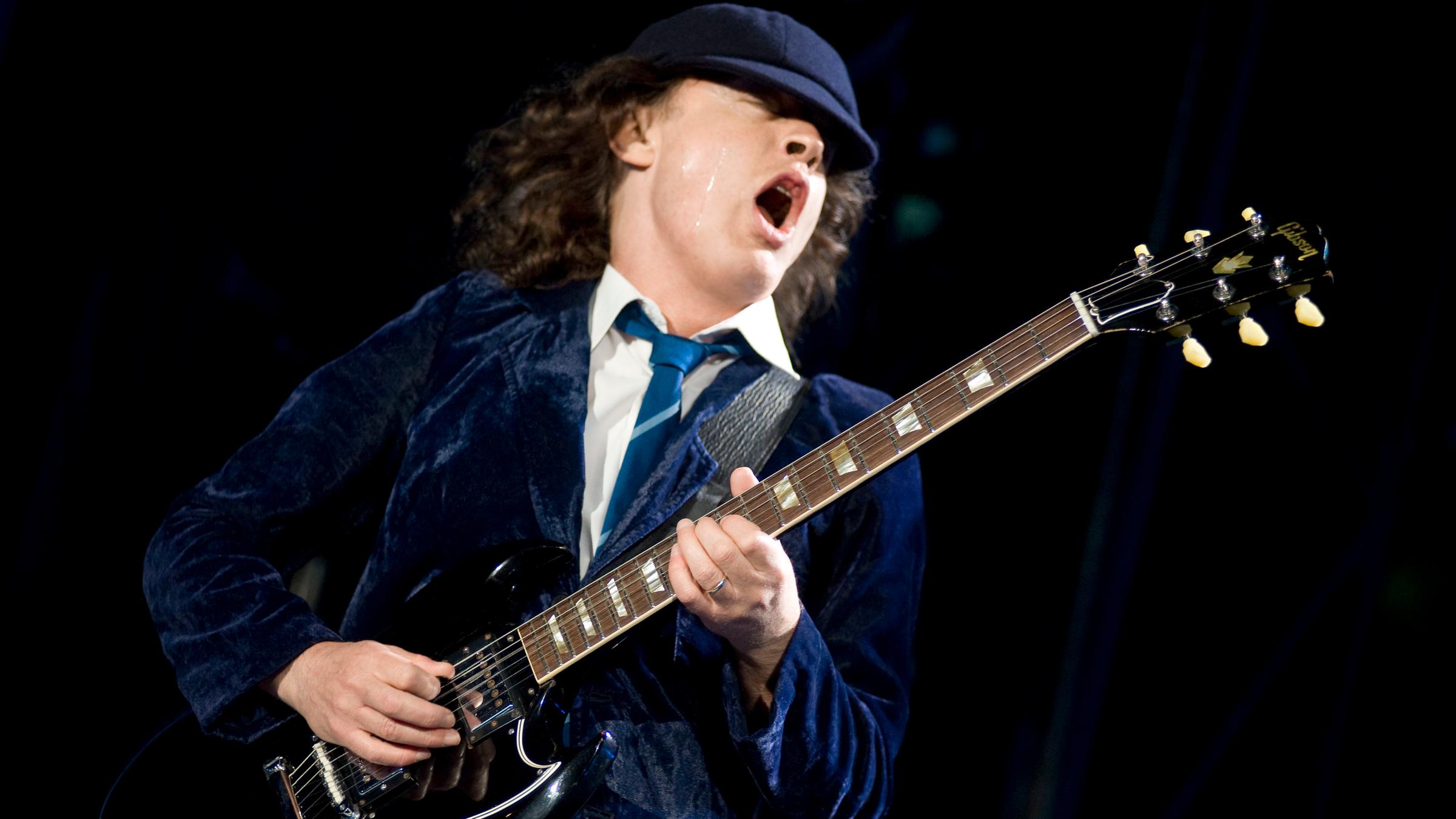 "Pop Around the Clock – Summer Edition: AC/DC: Live at Donington": Ein Mann mit dunkelblauem Samtsakko, Krawatte und weißem Hemd spielt auf einer E-Gitarre und singt mit geschlossenen Augen und weit geöffnetem Mund.