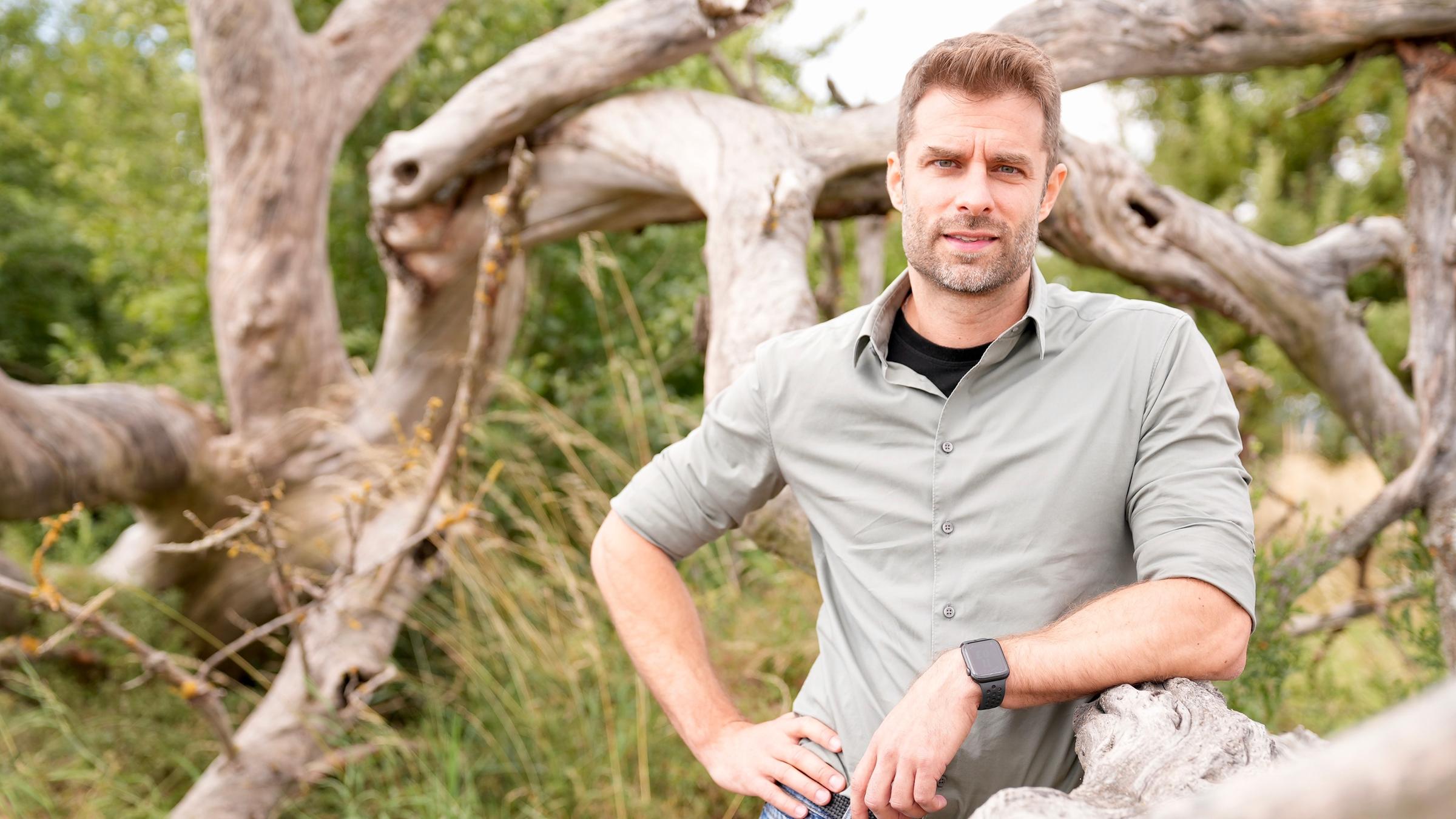 "ZDFzeit: Artensterben in Deutschland - die Fakten mit Kai Kupferschmidt": Kai Kupferschmidt lehnt rechts an einem Baumstamm, im Hintergrund weitere Baumstammäste und hohes Gras.