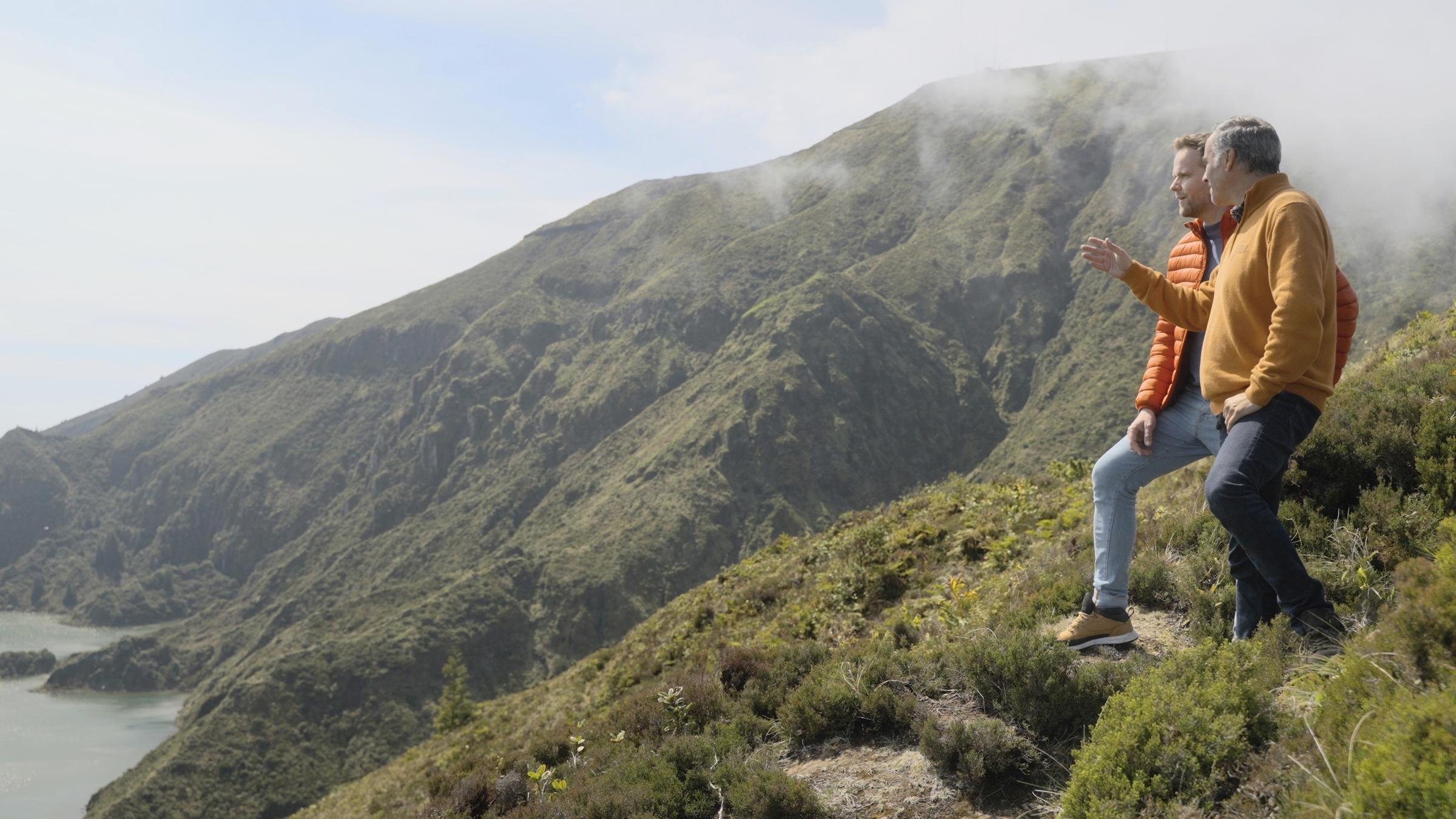 "Heiter bis sonnig – Wo unser Sommer gemacht wird": Benjamin Stöwe steht mit Inselführer Pedro Ferreira auf einer Anhöhe am Meeresufer. Die beiden schauen in die gleiche Richtung.