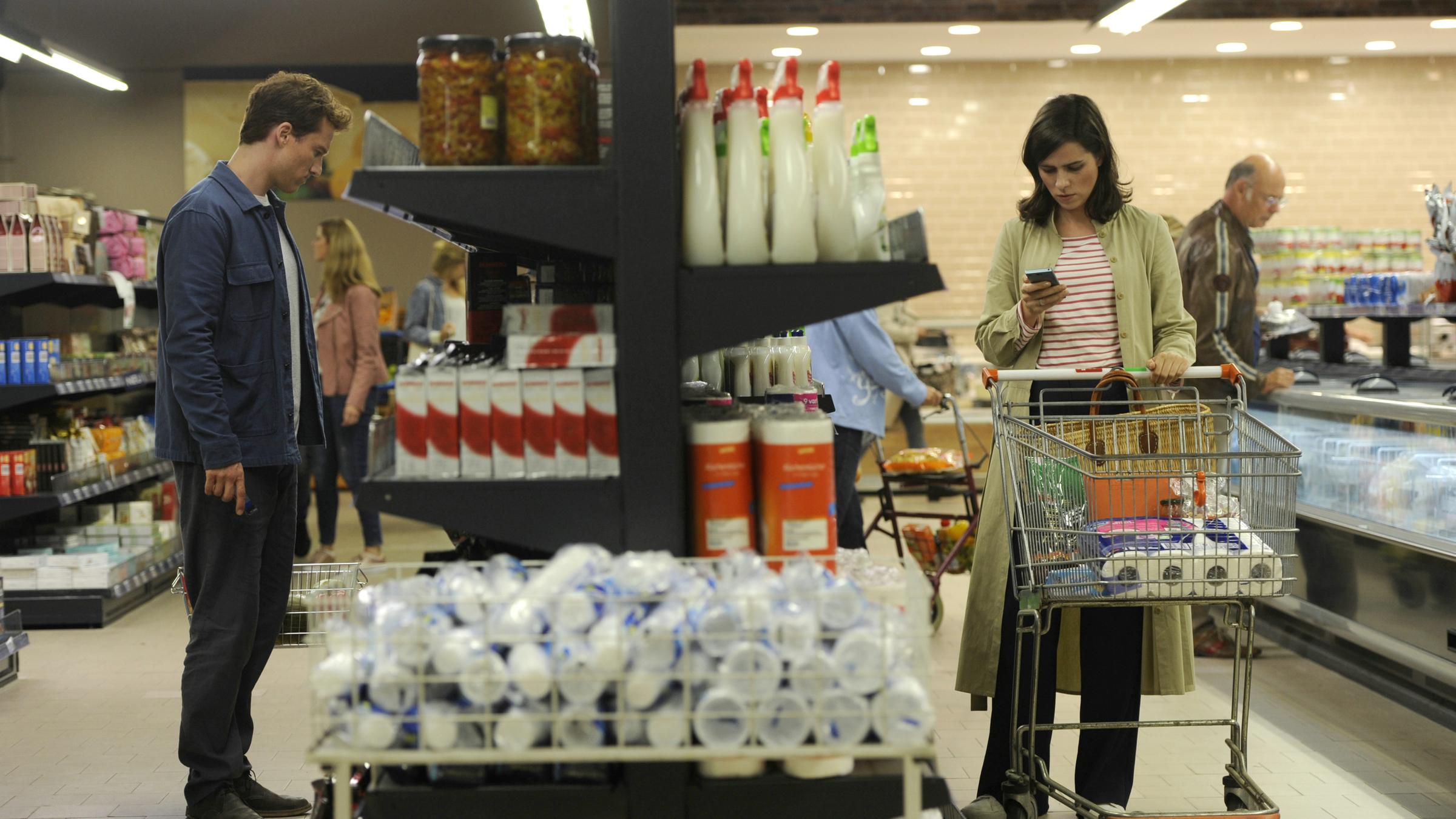 "Gut gegen Nordwind": Emma Rothner (Nora Tschirner) und Leo Leike (Alexander Fehling) stehen im Supermarkt auf der jeweils anderen Seite eines Regals. Emma schiebt einen Einkaufswagen und schaut auf ihr Handy, während Leo sich die Waren im Regal ansieht.
