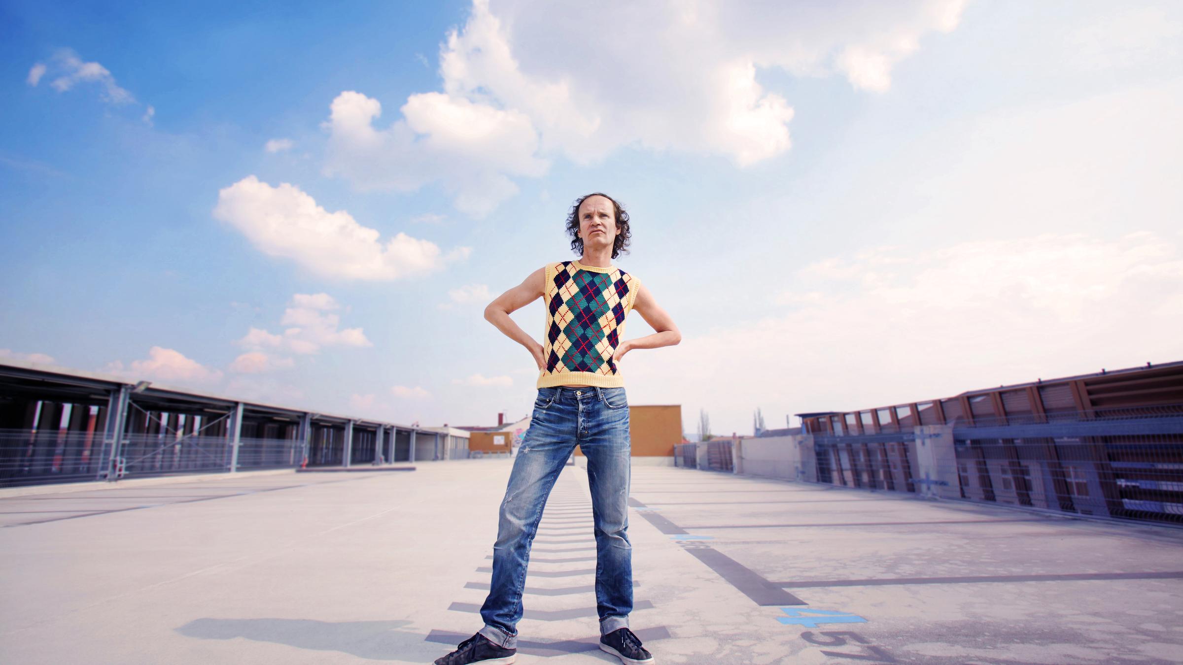 "Comedy for Future": Olaf Schubert steht breitbeinig auf einem großen asphaltierten Platz. Im Hintergrund leicht bewölkter Himmel.