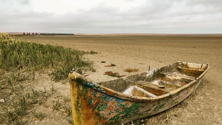 "Dürre in Europa - Die Katastrophe ist hausgemacht": Altes Boot auf trockenem, weitflächigem Sandboden. Zum Teil vertrocknete Halme und Gräser auf der linken Seite des Bildes.