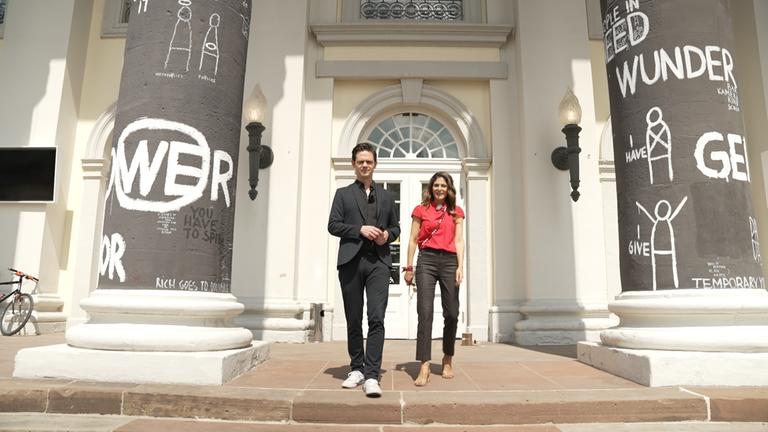 "aspekte: documenta 15: "Lumbung" oder die Kunst in Kassel": Die "aspekte"-Moderatoren Katty Salié und Jo Schück auf der documenta 15 in Kassel. Sie stehen auf der Eingangstreppe zu einem weiß gestrichenen Haus.