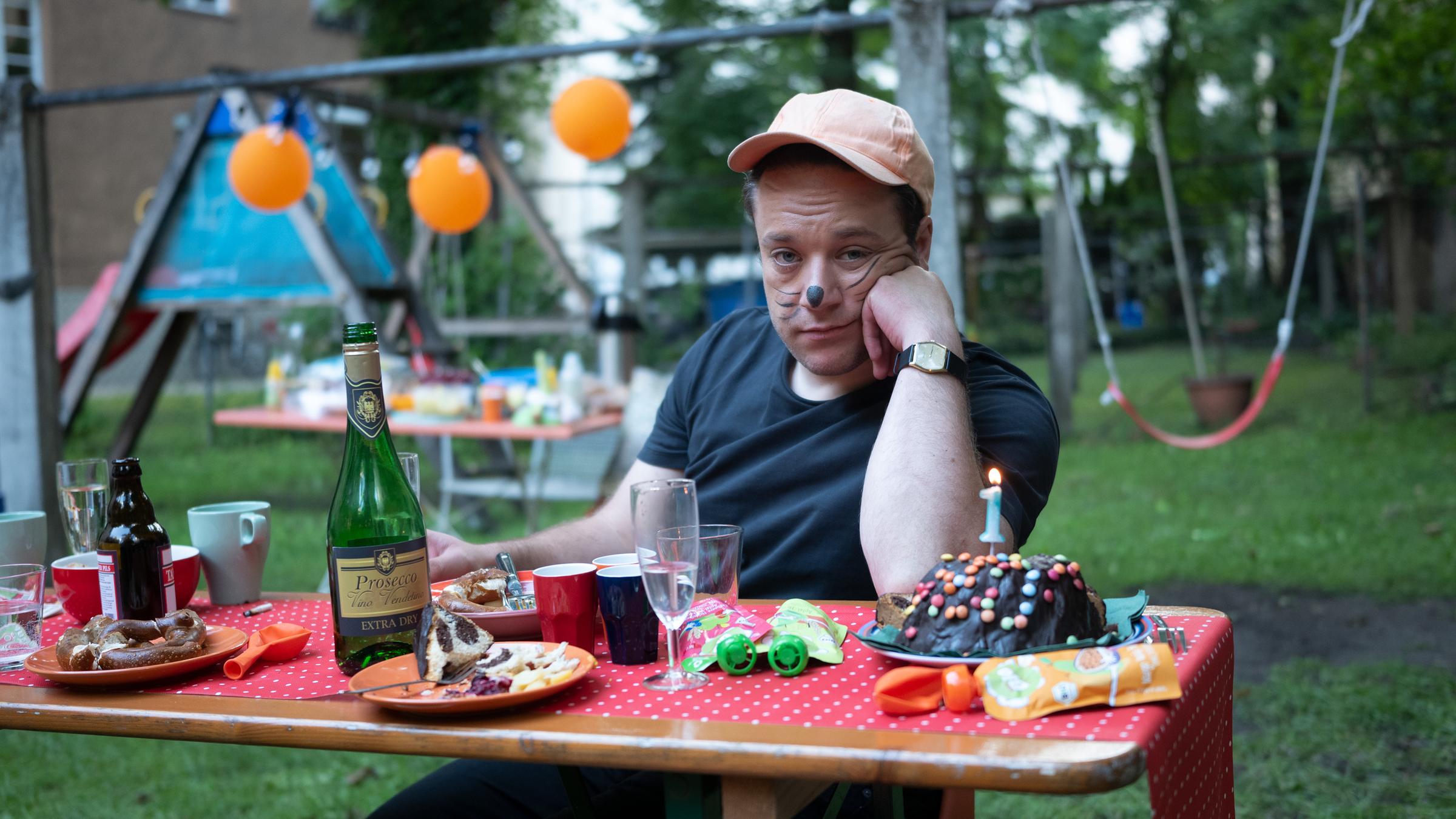 "FETT UND FETT - So kompliziert": Jaksch (Jakob Schreier) sitzt an einem mit Kuchen und Getränken beladenen Biertisch. Er stütz seinen Kopf mit seinem linken Arm ab, ist im Gesicht bemalt und sieht stirnrunzelnd in die Kamera.