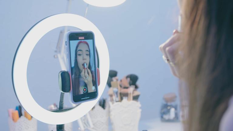 "This is TikTok - Stars und Hype in Hochkant": Die 17-jährige Hasti sieht sich in der Innenkamera ihres Handys an und schminkt sich dabei die Lippen. Das Handy ist an einem Ringlicht befestigt und auf einem Tisch befinden sich mehrere Make-up-Pinsel.