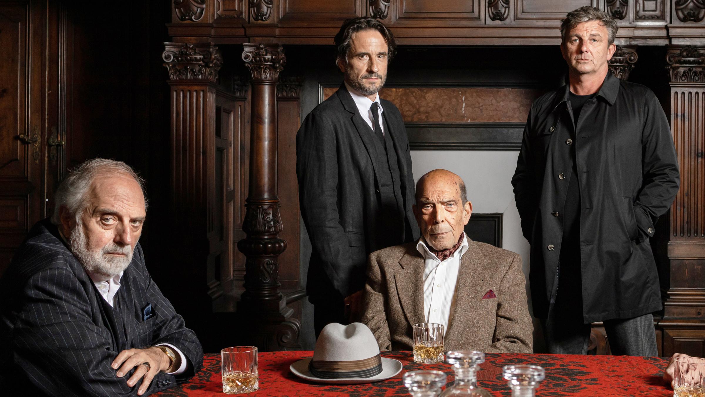 "Der Feind meines Feindes": Pietro Connagi (Alessandro Bressanello) und Toto Conte (Orso Maria Guerrini) sitzen an einem rot-schwarzen Tisch und blicken ernst in die Kamera. Auf dem Tisch stehen zwei halbvolle Whiskeygläser und zwischen diesen liegt ein grauer Hut. Hinter Toto Conte stehen Gabriel Morales (Oliver Mommsen) und Klaus Burg (Hans Sigl) nebeneinander und sehen ebenfalls in die Kamera. Klaus Burger trägt einen schwarzen Anorak, die anderen Männer auf dem Bild sind mit Anzügen bekleidet.