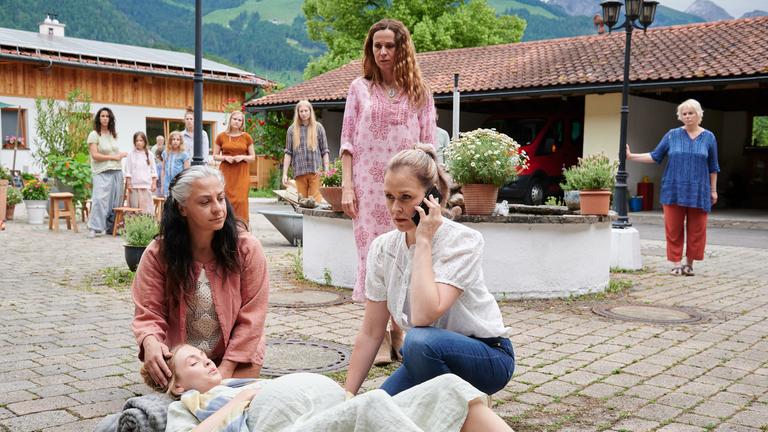 "Lena Lorenz - Freiheit": Auf dem Pflasterboden eines Innenhofes liegt die hochschwangere Lilly Waldhauser (Teresa Klamert). Tine Michaelis (Göksen Güntel) stützt ihr den Kopf. Bei ihr kniet Lena Lorenz (Judith Hoersch), die gerade telefoniert. Hinter ihnen steht Marthe Gerber (Doris Schretzmayer). In Entfernung beobachten einige Bewohner der Landkommune das Geschehen.