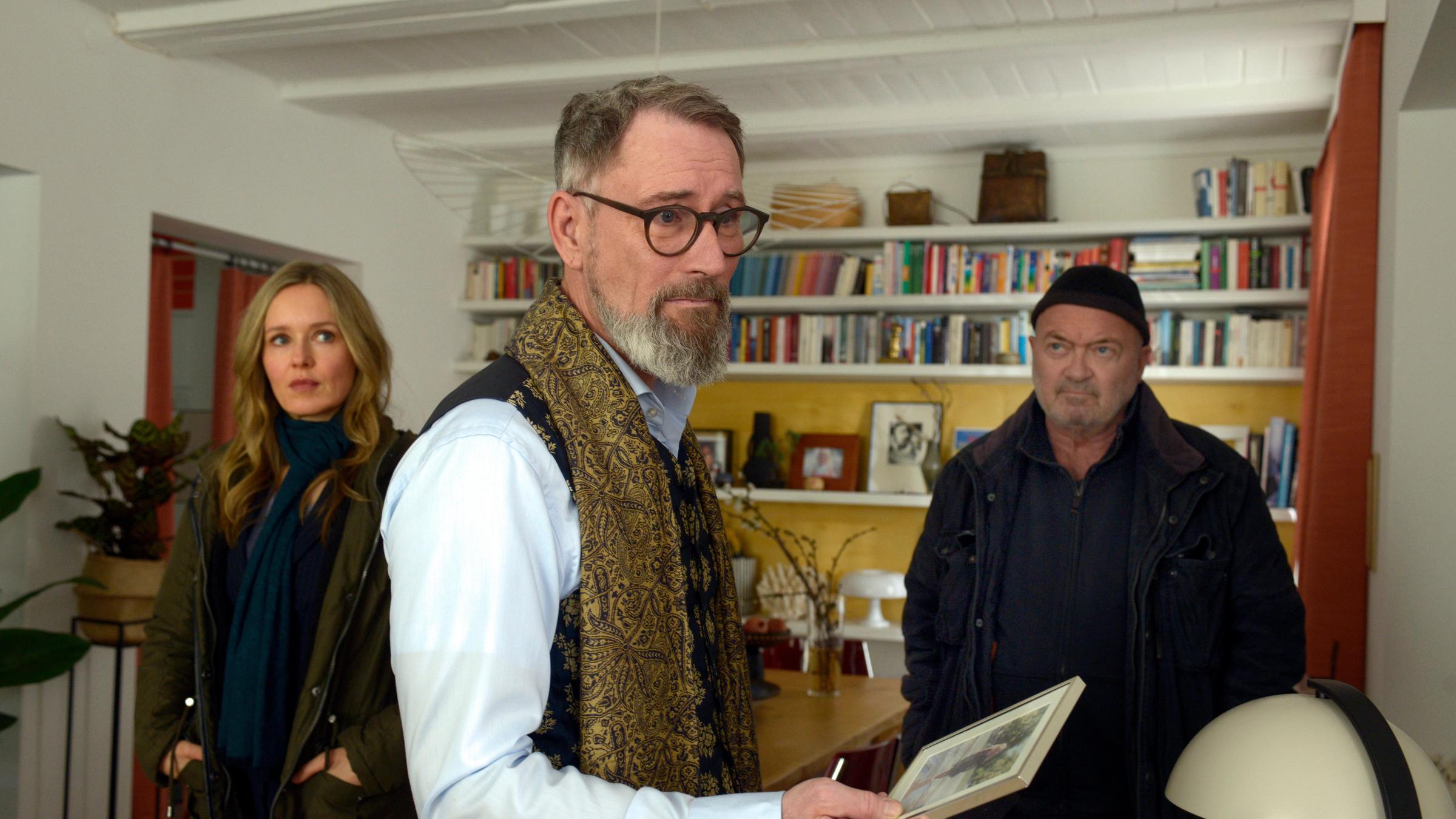 "Ein starkes Team – Abgestürzt": In einem Wohnzimmer mit einer Bücherwand: Rolf Christiansen (Heikko Deutschmann) steht im Vordergrund mit einem Bild in der Hand und blickt besorgt nach vorne. Hinter ihm stehen Linett Wachow (Stefanie Stappenbeck) und Otto Garber (Florian Martens) und schauen ihn an.
