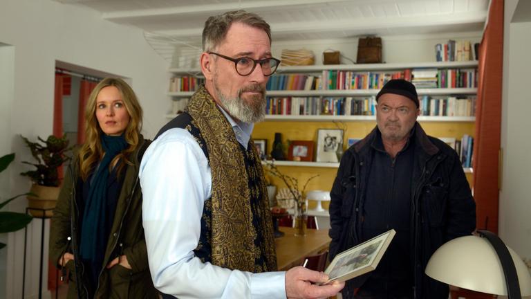 "Ein starkes Team – Abgestürzt": In einem Wohnzimmer mit einer Bücherwand: Rolf Christiansen (Heikko Deutschmann) steht im Vordergrund mit einem Bild in der Hand und blickt besorgt nach vorne. Hinter ihm stehen Linett Wachow (Stefanie Stappenbeck) und Otto Garber (Florian Martens) und schauen ihn an.