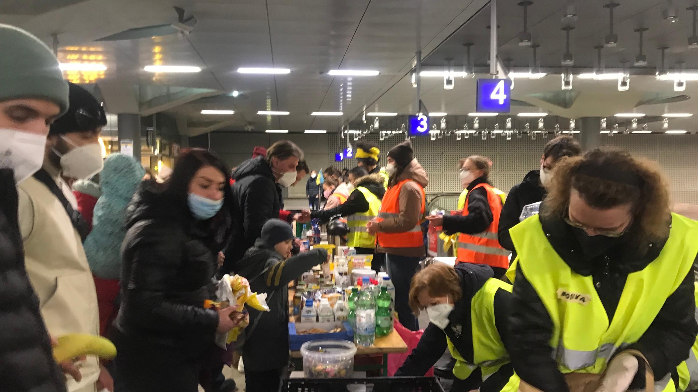 "ZDF.reportage: Ukraine-Krieg erschüttert Deutschland": Freiwillige Helferinnen in gelben und orangen Warnwesten verteilen am Hauptbahnhof Berlin gespendete Lebensmittel an ankommende Geflüchtete aus der Ukraine.