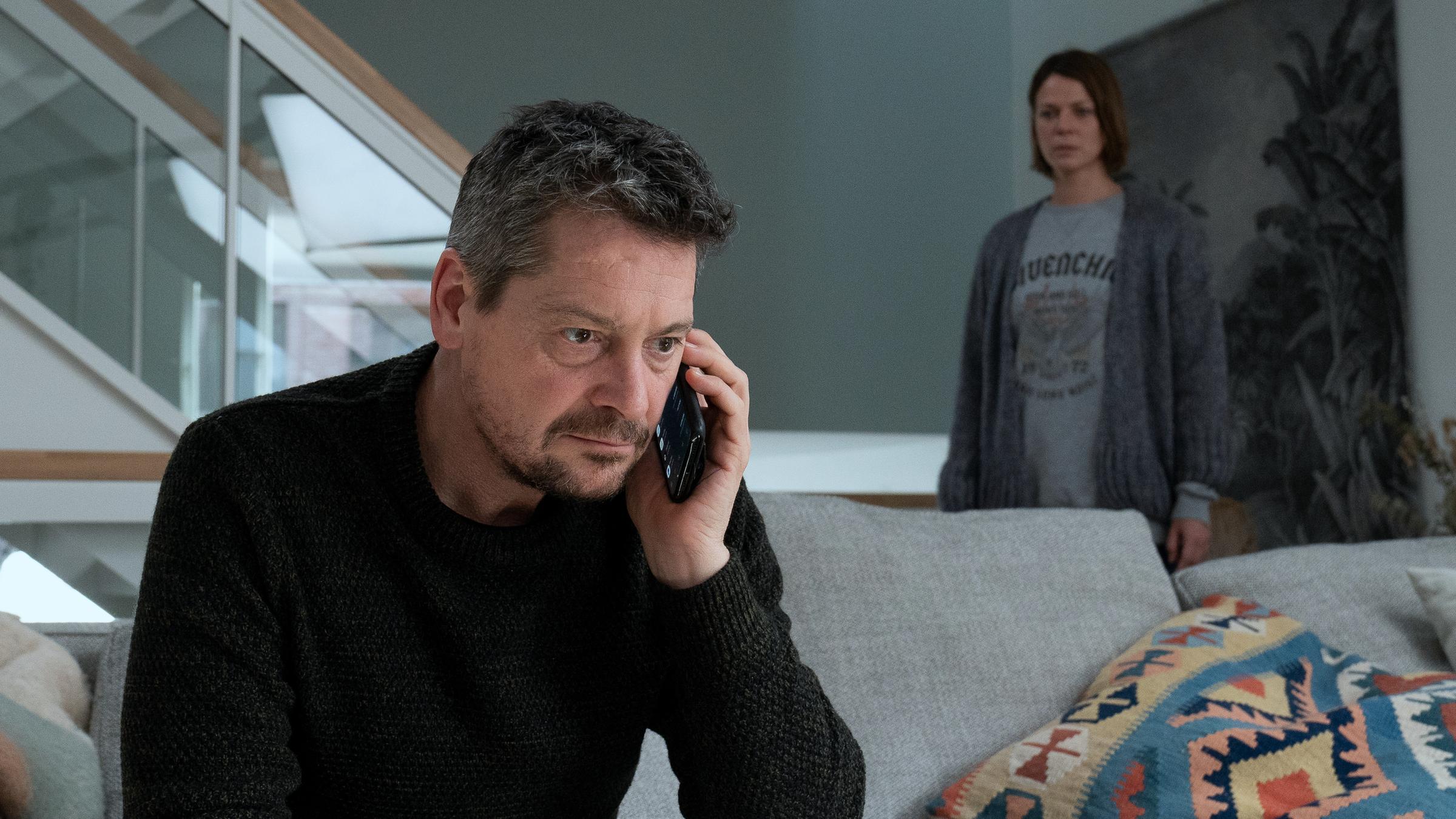 "Alles auf Rot": Kessel (Fritz Karl) sitzt in einem Wohnzimmer auf einem grauen Sofa und telefoniert. Claire (Jessica Schwarz) steht schräg hinter ihm an einer Treppe und belauscht das Telefonat.