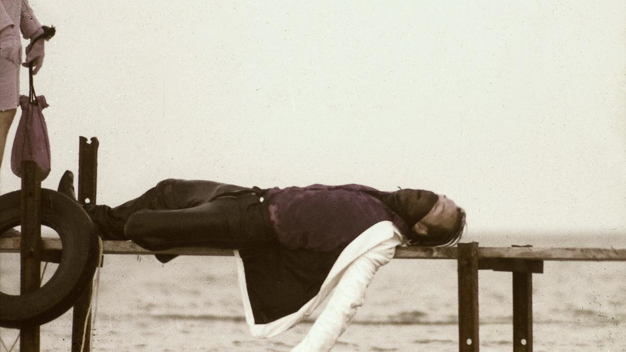 "Die Bettwurst": Steve liegt tot auf einem Steg. Ein Arm hängt herunter, die Augen sind geschlossen. Links neben im am Bildrand ist Luzi Kryn mit Handtasche zu erkennen.