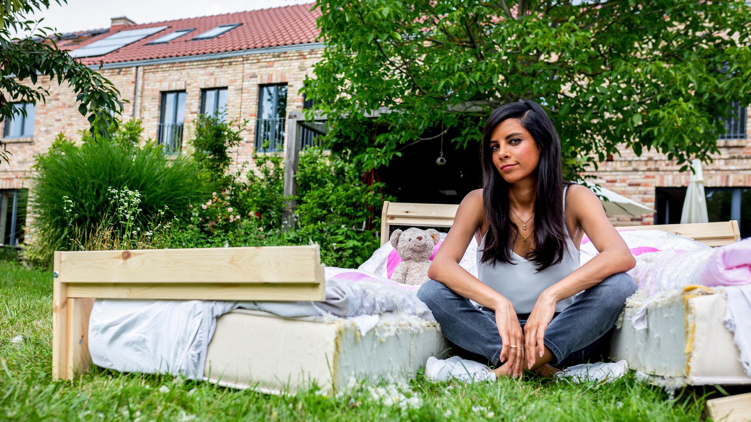 "Trennungskinder - Wenn Eltern auseinandergehen (1)": Moderatorin Collien Ulmen-Fernandes sitzt in einem Garten im Schneidersitz zwischen den beiden Hälften eines auseinandergesägten Ehebetts. Auf dem Bett ein Teddybär. Im Hintergrund Häuser, Büsche und Bäume.