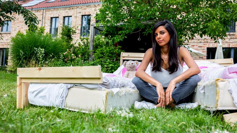 "Trennungskinder - Wenn Eltern auseinandergehen (1)": Moderatorin Collien Ulmen-Fernandes sitzt in einem Garten im Schneidersitz zwischen den beiden Hälften eines auseinandergesägten Ehebetts. Auf dem Bett ein Teddybär. Im Hintergrund Häuser, Büsche und Bäume.