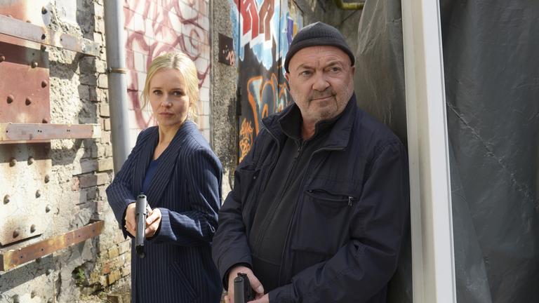 "Ein starkes Team - Die letzte Reise": Linett Wachow (Stefanie Stappenbeck) und Otto Garber (Florian Martens) stehen im Gang zwischen einer Plane und einer mit Graffiti bemalten Wand und halten jeweils eine Pistole schräg nach unten vor sich.