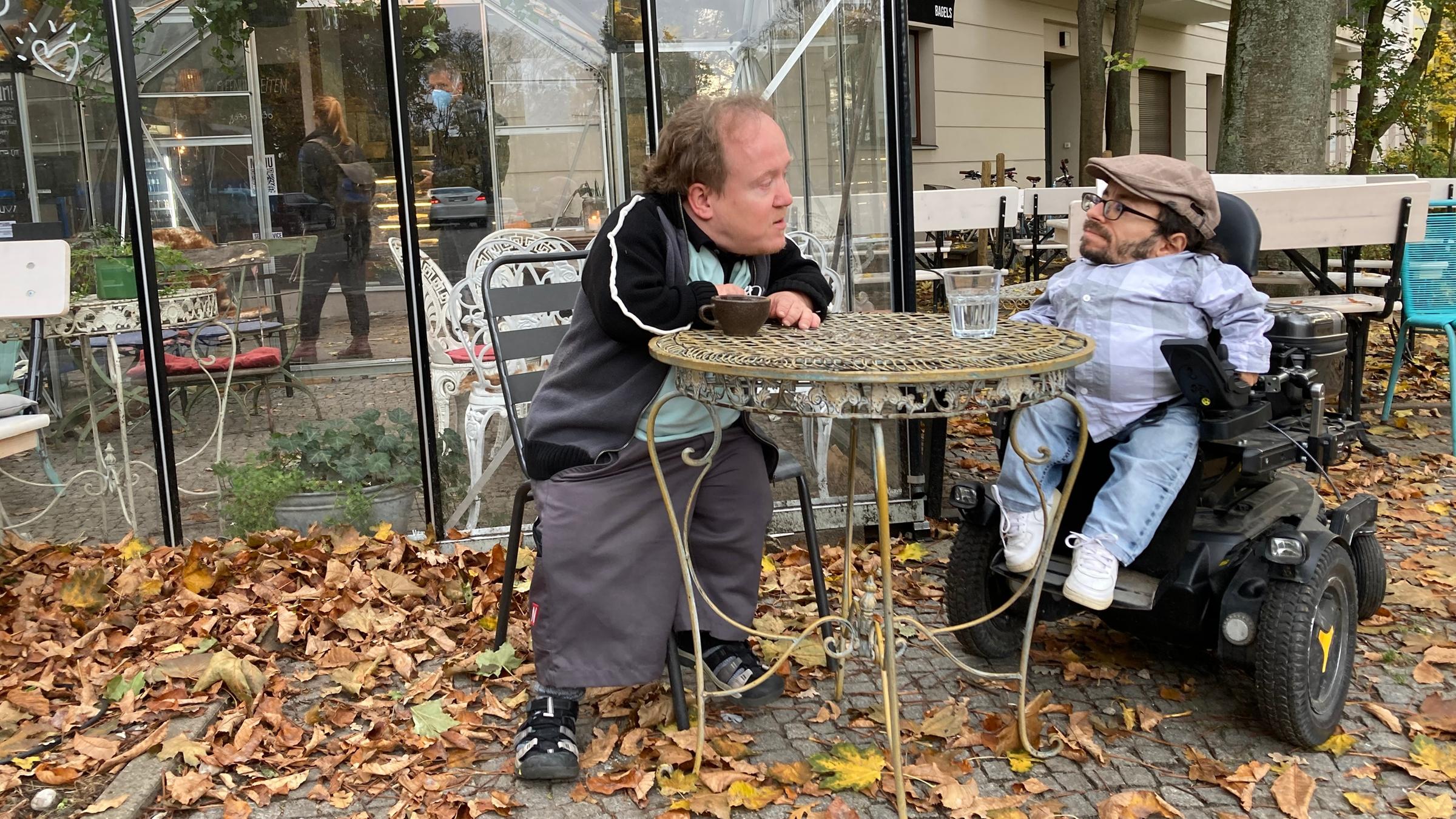 "Geliebt, geduldet, getötet - Die Geschichte von Menschen mit Behinderungen": Martin Müller steht an einem Tisch, draußen vor einem Café. Der Boden ist bedeckt mit herbstlich braunen Blättern. Neben ihm sitzt Raul Krauthausen mit Schiebermütze im Rollstuhl.