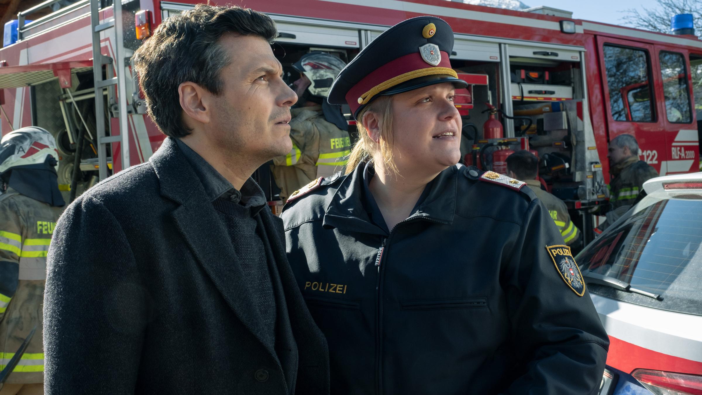 "Flammenmädchen": Martin Merana (Manuel Rubey) und Franziska Heilmayr (Stefanie Reinsperger) stehen nebeneinander vor einem Löschfahrzeug und schauen auf etwas außerhalb des Bildes.