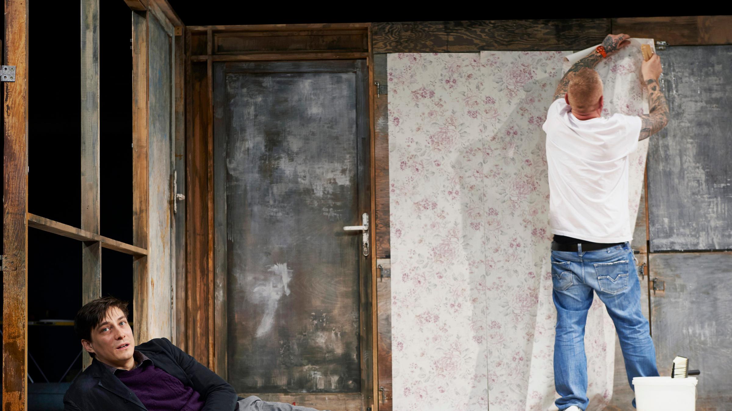 "Starke Stücke: Ein Mann seiner Klasse": Ein junger Mann mit dunklen Haaren liegt auf dem Boden, aufgestützt auf seinen rechten Arm. Er trägt eine hellgraue Hose, ein dunkelgraues Sakko und einen lila Pullover. Im Hintergrund steht mit dem Rücken zum Publikum ein etwas älterer Mann, mit kurzgeschorenen Haaren, die Arme sind von Tattoos bedeckt, er trägt ein weißes T-Shirt und eine helle Jeans, er tapeziert die Rückwand der Kulisse.