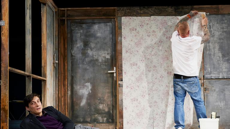 "Starke Stücke: Ein Mann seiner Klasse": Ein junger Mann mit dunklen Haaren liegt auf dem Boden, aufgestützt auf seinen rechten Arm. Er trägt eine hellgraue Hose, ein dunkelgraues Sakko und einen lila Pullover. Im Hintergrund steht mit dem Rücken zum Publikum ein etwas älterer Mann, mit kurzgeschorenen Haaren, die Arme sind von Tattoos bedeckt, er trägt ein weißes T-Shirt und eine helle Jeans, er tapeziert die Rückwand der Kulisse.