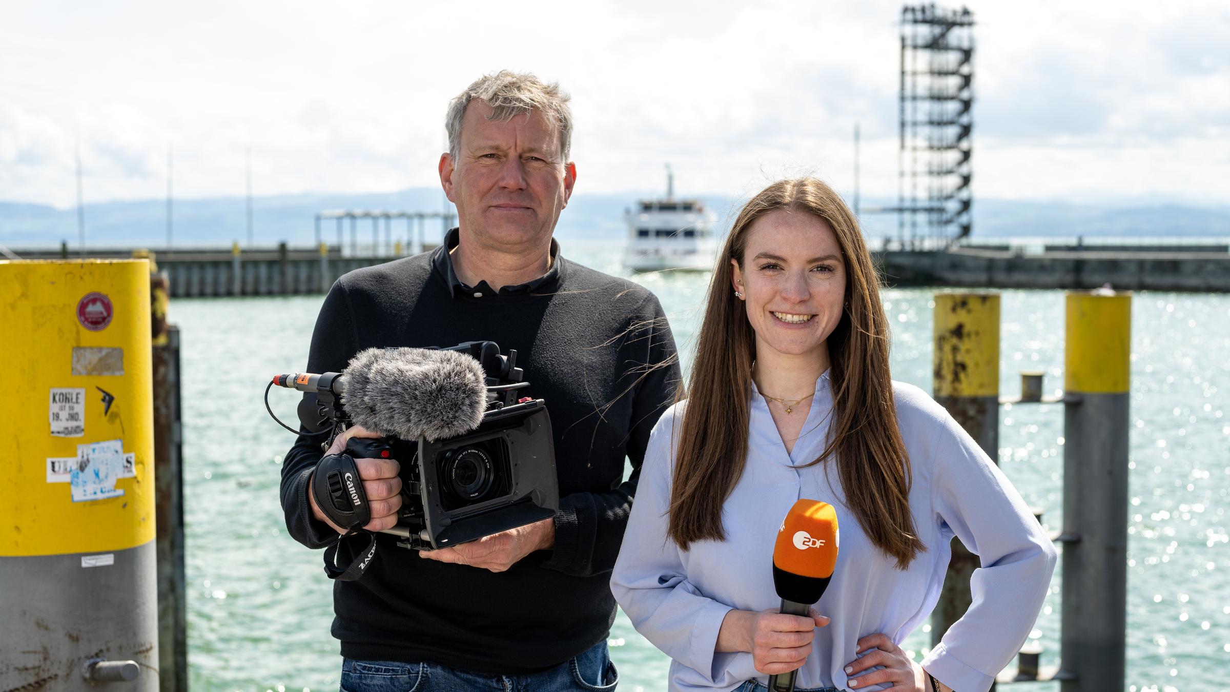 ZDF in Friedrichshafen (20. ZDF-Bürgerprojekt) Mai 2022:  Kameramann Manuel Brückl und Reporterin Anna Gürth stehen nebeneinander vor dem Schiffsanleger in Friedrichshafen und blicken in die Kamera.
