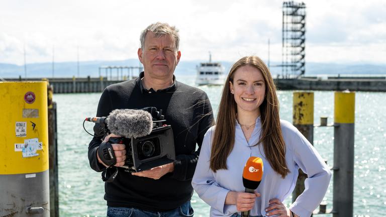 ZDF in Friedrichshafen (20. ZDF-Bürgerprojekt) Mai 2022:  Kameramann Manuel Brückl und Reporterin Anna Gürth stehen nebeneinander vor dem Schiffsanleger in Friedrichshafen und blicken in die Kamera.