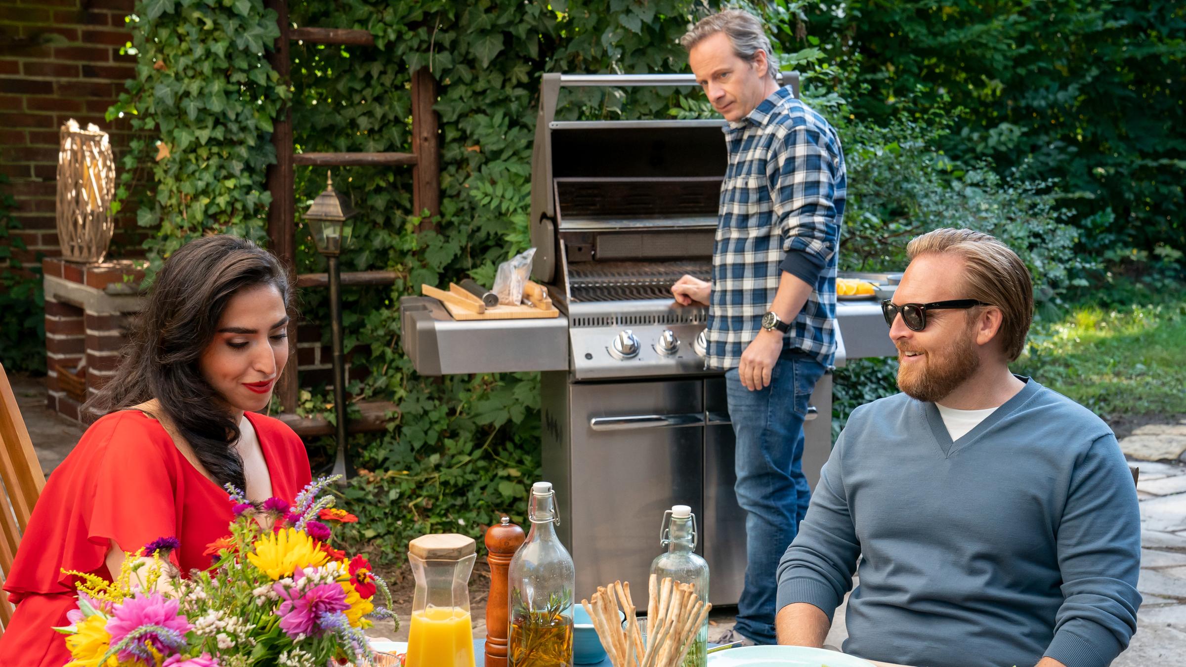 "Der Spalter": Dila (Susana AbdulMajid) und Lars (Axel Stein) sitzen auf eine Terrasse an einem gedeckten Tisch und unterhalten sich. Oliver (Fabian Busch) steht im Hintergrund an einem Grill und beobachtet die Beiden.
