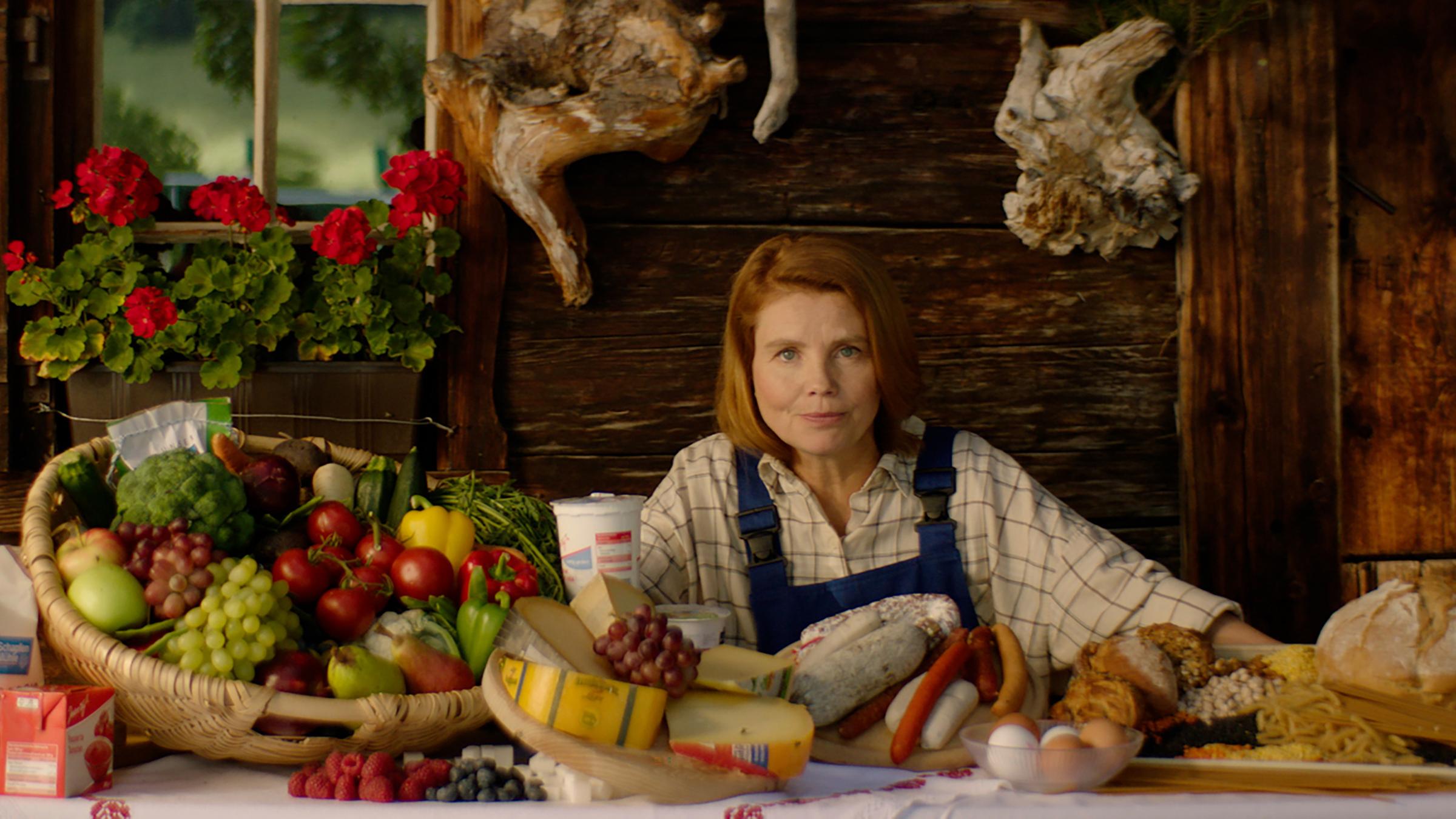 "ZDFzeit: Die Wahrheit über unsere Landwirtschaft - von Geldgier, Investoren, und Getreide": Annette Frier mit einer Latzhose sitzt an einem gedeckten Tisch mit Obst, Gemüse und Käse.